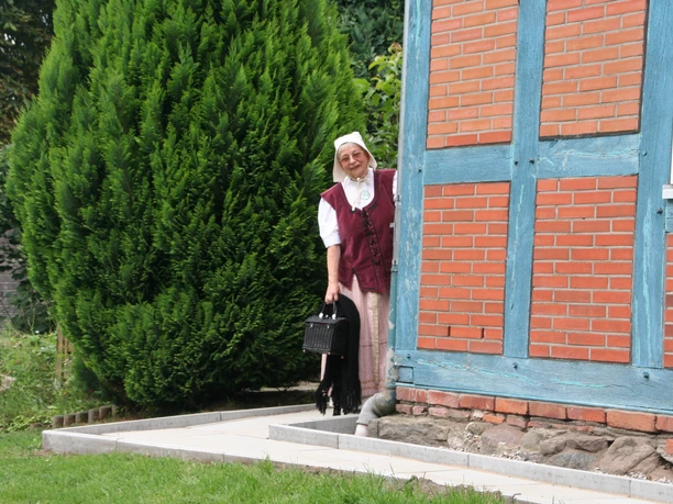 Magd Berta Eine Frau in historischer Tracht blickt lächelnd hinter einer Fachwerkfassade hervor, umgeben von Grün.