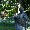 Steinskulptur eines Menschen mit Hut, der eine Zeitung hält, umgeben von Grün und Bäumen.