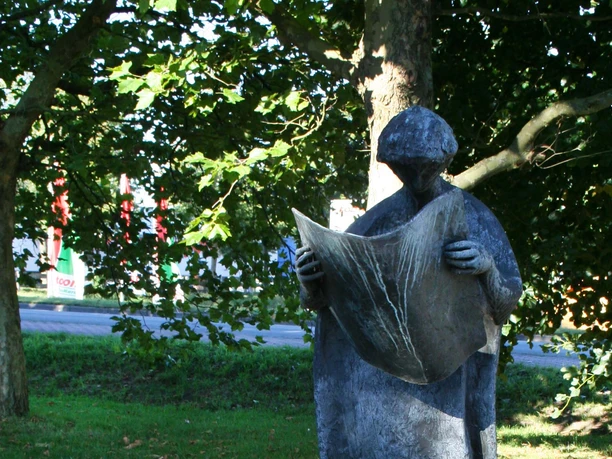 Steinskulptur eines Menschen mit Hut, der eine Zeitung hält, umgeben von Grün und Bäumen.