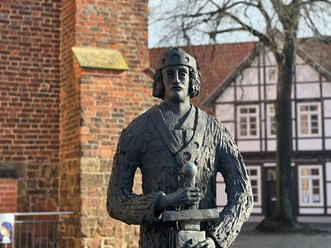 Skulptur von Widukind vor einem Fachwerkhaus, hält Schwert und ist mit mittelalterlicher Kleidung dargestellt.