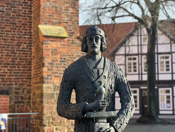 Skulptur von Widukind vor einem Fachwerkhaus, hält Schwert und ist mit mittelalterlicher Kleidung dargestellt.