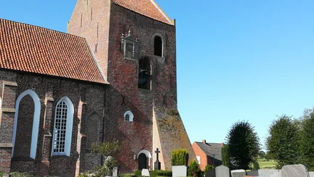 Kirche Groothusen Backsteinkirche mit hohem Turm und rotem Ziegeldach vor blauem Himmel, umgeben von Grabsteinen.