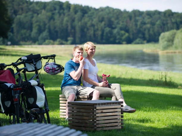 Picknick an der Weser Ein Paar genießt ein Picknick am Flussufer der Weser, umgeben von grüner Natur und Fahrrädern.