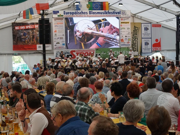 Large tent with lots of people, brass band theater on stage, international flags.
