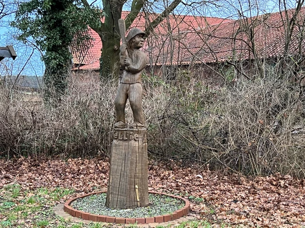 A wooden statue of a rounders player named Wellie, surrounded by winter trees and leaves.