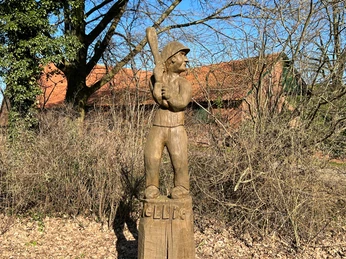 Schlagballspieler Wellie Holzstatue eines Schlagballspielers in historischer Sportausrüstung auf einem Sockel im Grünen.