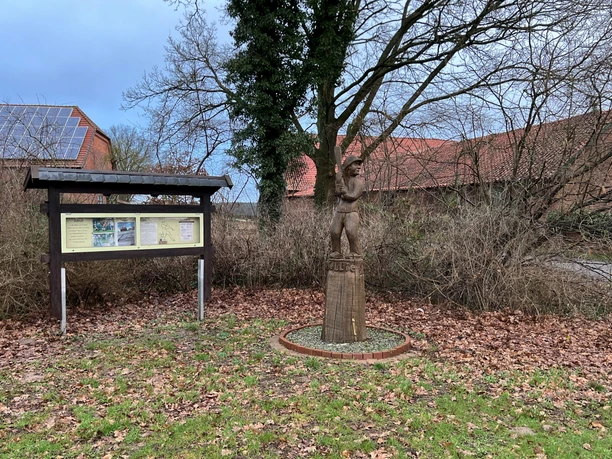 Eine Statue eines Schlagballspielers aus Holz steht auf einem Sockel vor einer Informations-Tafel und Bäumen.