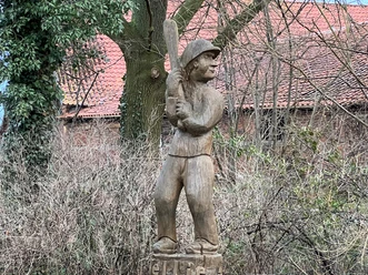 Holzskulptur eines Schlagballspielers im Wald, zeigt feste Haltung und traditionellen Spielstil.