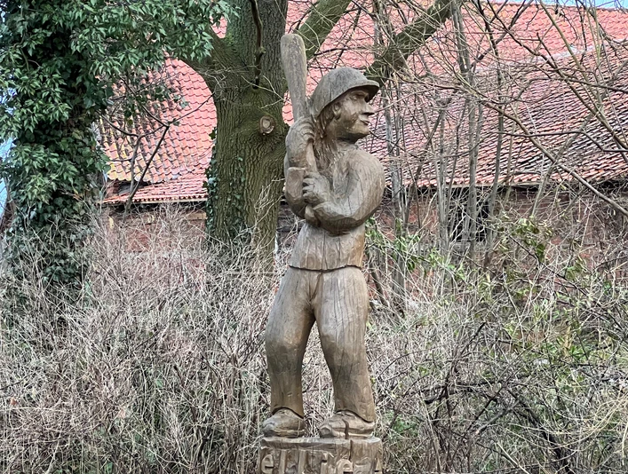 Schlagballspieler Wellie Holzskulptur eines Schlagballspielers im Wald, zeigt feste Haltung und traditionellen Spielstil.