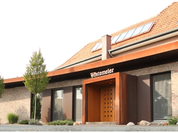 Modernes Gebäude mit Backsteinfassade, großem Holzportal und umgebenden Bäumen im Vordergrund.