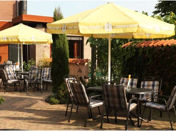 Ein gemütlicher Biergarten mit karierten Stühlen und gelben Warsteiner-Sonnenschirmen an einem sonnigen Tag.