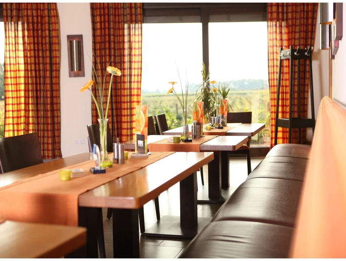 Lichtdurchfluteter Raum mit Holztischen, orange-weißen Vorhängen und Blick auf eine grüne Landschaft.