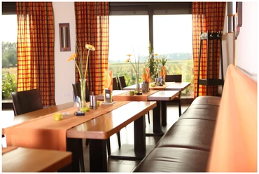 Gaststätte Wöstemeier_Restaurant Lichtdurchfluteter Raum mit Holztischen, orange-weißen Vorhängen und Blick auf eine grüne Landschaft.