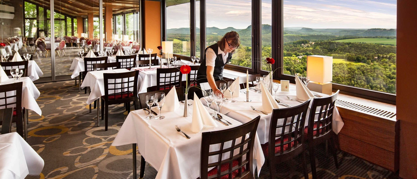 Ein Restaurant mit gedeckten Tischen, weißen Tischdecken und roten Blumen, ein Kellner richtet Besteck; große Fenster bieten eine Aussicht auf grüne Hügel.