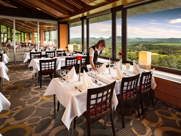 Panoramarestaurant Bastei Ein Restaurant mit gedeckten Tischen, weißen Tischdecken und roten Blumen, ein Kellner richtet Besteck; große Fenster bieten eine Aussicht auf grüne Hügel.