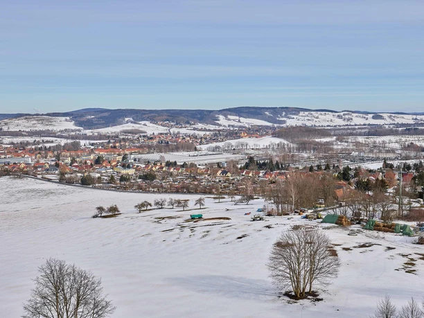 View from the Götzinger Höhe tower Snowy landscape with a village in the background, surrounded by hills; colorful houses and bare trees under a clear blue sky.