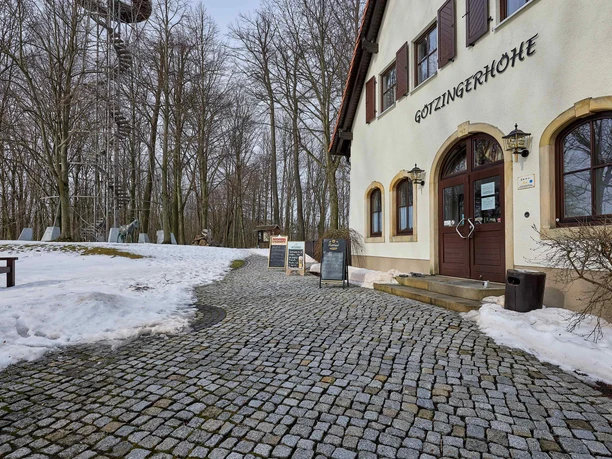 A building with the inscription "Götzingerhöhe" next to a snow-covered path; in the background a tower and bare trees.