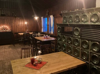 Innenraum Bergwirtschaft Papststein Gemütlicher Raum mit Holzwänden, Tischen und Stühlen, dekoriert mit Kerzen und Weihnachtskugeln; ein grüner Kachelofen steht im Vordergrund.Cozy room with wooden walls, tables and chairs, decorated with candles and Christmas baubles; a green tiled stove stands in the foreground.Útulná místnost s dřevěnými stěnami, stoly a židlemi, vyzdobená svíčkami a vánočními ozdobami; v popředí stojí zelená kachlová kamna.Przytulny pokój z drewnianymi ścianami, stołami i krzesłami, udekorowany świecami i świątecznymi bombkami; na pierwszym planie stoi zielony piec kaflowy.Gezellige kamer met houten muren, tafels en stoelen, versierd met kaarsen en kerstballen; een groen betegelde kachel staat op de voorgrond.Accogliente sala con pareti in legno, tavoli e sedie, decorata con candele e decorazioni natalizie; in primo piano una stufa in maiolica verde.