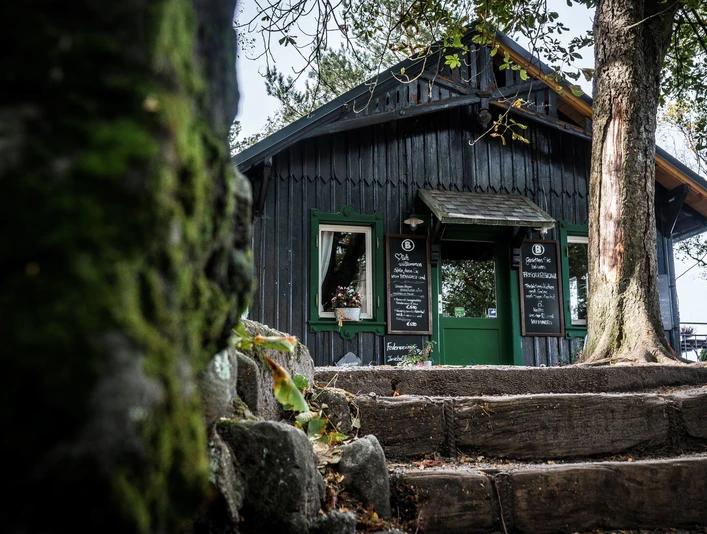 Holzhütte mit grünen Fensterrahmen und Tür, umgeben von Bäumen; zwei Tafeln mit Menü stehen neben dem Eingang, herbstliche Atmosphäre.Wooden hut with green window frames and door, surrounded by trees; two tables with menu next to the entrance, autumnal atmosphere.Dřevěná chata se zelenými okenními rámy a dveřmi, obklopená stromy; dvě tabule s jídelním lístkem vedle vchodu, podzimní atmosféra.Drewniana chata z zielonymi ramami okiennymi i drzwiami, otoczona drzewami; dwa stoliki z menu obok wejścia, jesienna atmosfera.Houten hut met groene kozijnen en deur, omringd door bomen; twee tafels met menu naast de ingang, herfstsfeer.Capanna in legno con infissi e porta verdi, circondata da alberi; due tavoli con menu accanto all'ingresso, atmosfera autunnale.