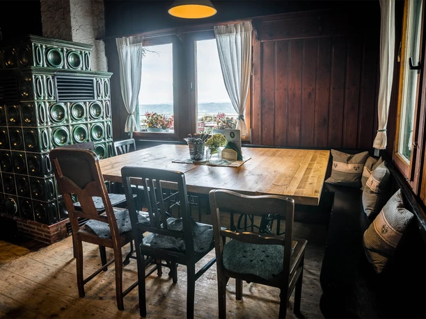 Gezellige houten hoek met grote tafel, groene tegelkachel, stoelen en kussens, uitzicht op bloeiende planten en landschap door het raam.