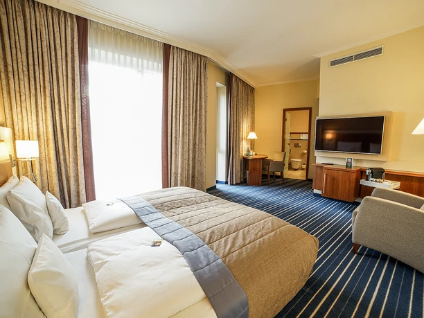 Elegantes Hotelzimmer mit Doppelbett, gemütlichem Sessel, Schreibtisch und großem Flachbildfernseher.