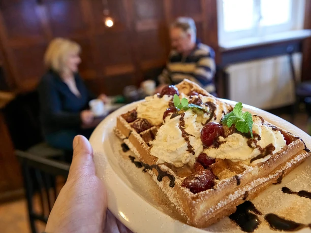 Waffle with cream, cherries and chocolate sauce on a plate, in the background a couple in a cozy café.
