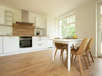 Heller Ess- und Wohnbereich mit Holzfußboden, moderner Küche und großen Fenstern mit Blick ins Grüne.