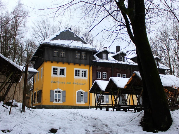 Bergwirtschaft Kuhstall Gelbes Gasthaus mit grauen Fensterläden im verschneiten Wald, Holztische im Vordergrund, winterliche Stimmung.