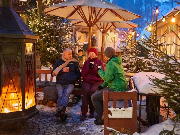 Three people sit around a fireplace in a wintry atmosphere, surrounded by snow-covered fir trees and colorful lights, drinking hot drinks.
