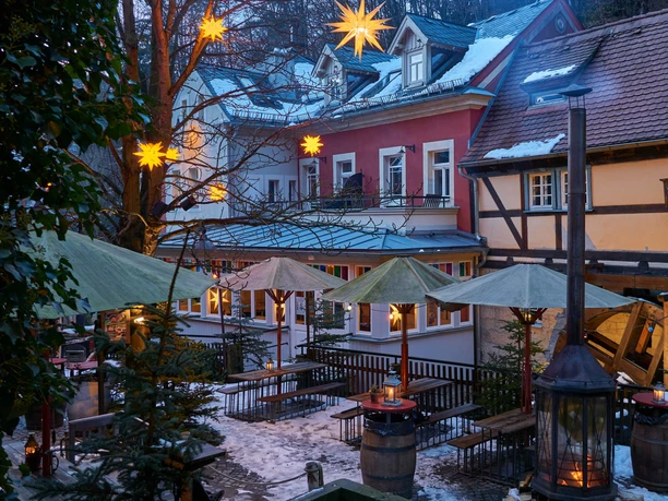 Snow-covered inner courtyard with illuminated stars on trees, wooden benches, green parasols and lanterns, surrounded by half-timbered houses.