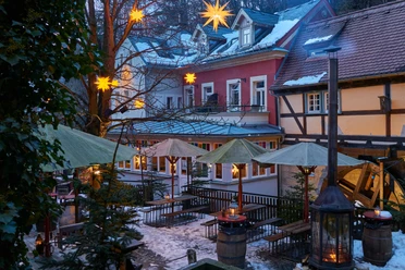 Winterdorf Schmilka Mühlenhof Verschneiter Innenhof mit beleuchteten Sternen an Bäumen, Holzbänken, grünen Sonnenschirmen und Laternen, umgeben von Fachwerkhäusern.