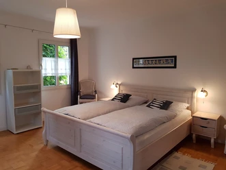 Helles Schlafzimmer mit Doppelbett, weißen Möbeln, Fenster, Vorhängen, Lampe und dekorativem Bild.