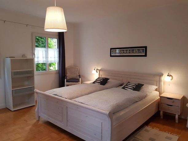 Schlafzimmer.jpg Helles Schlafzimmer mit Doppelbett, weißen Möbeln, Fenster, Vorhängen, Lampe und dekorativem Bild.