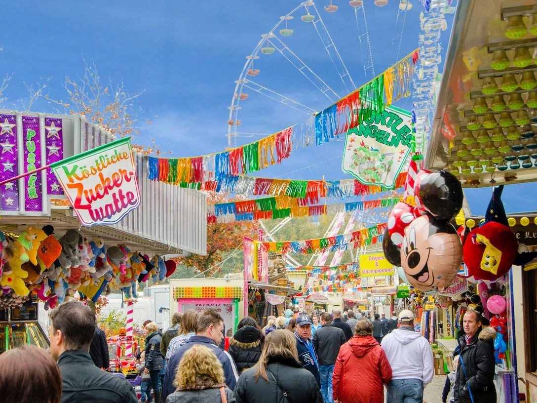 kirmes_meppen.jpg Menschen schlendern zwischen bunten Buden auf einer belebten Kirmes mit Riesenrad und Luftballons.