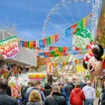 kirmes_meppen.jpg Menschen schlendern zwischen bunten Buden auf einer belebten Kirmes mit Riesenrad und Luftballons.
