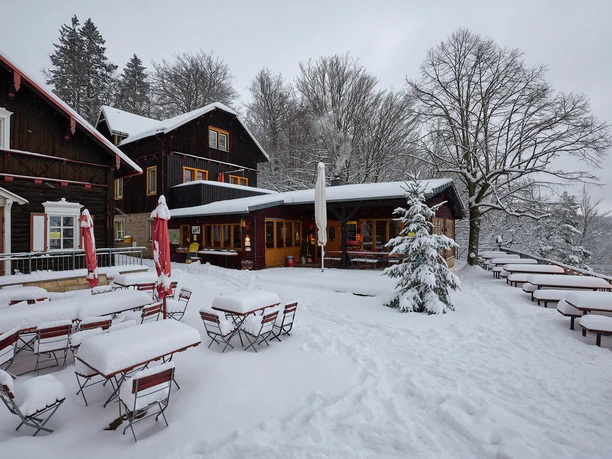 Besneeuwd terras van een houten huis met rode parasols en tafels, omringd door besneeuwde bomen, winterse sfeer.