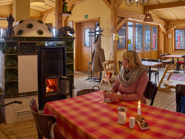 Een vrouw zit aan een tafel met een rood geruit tafelkleed in een gezellige kamer, naast een brandende kachel, en houdt een kopje vast.