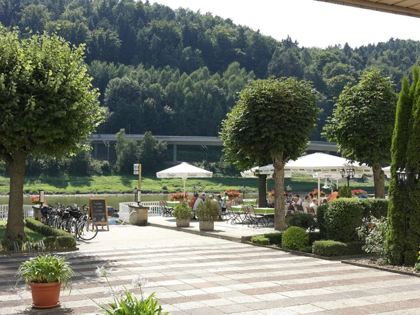 Terrasse Elbhotel Bad Schandau Gepflegte Terrasse mit Tischen und weißen Sonnenschirmen, umgeben von grünen Bäumen und Pflanzen, im Hintergrund ein bewaldeter Hügel.