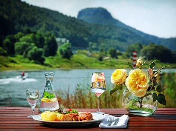 Elbhotel Bad Schandau Ein gedeckter Tisch mit einem Teller Essen, einer Karaffe Wasser und einem Glas Wein steht vor einer Flusslandschaft mit gelben Rosen im Vordergrund.A laid table with a plate of food, a carafe of water and a glass of wine stands in front of a river landscape with yellow roses in the foreground.Před říční krajinou se žlutými růžemi v popředí stojí prostřený stůl s talířem s jídlem, karafou s vodou a sklenicí vína.Nakryty stół z talerzem jedzenia, karafką wody i kieliszkiem wina stoi przed krajobrazem rzeki z żółtymi różami na pierwszym planie.Een gedekte tafel met een bord eten, een karaf water en een glas wijn staat voor een rivierlandschap met gele rozen op de voorgrond.Una tavola imbandita con un piatto di cibo, una caraffa d'acqua e un bicchiere di vino si trova davanti a un paesaggio fluviale con rose gialle in primo piano.