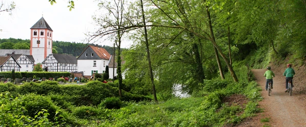 an der Dhünn mit Blick auf Odenthal Fahrradfahrer auf einem Waldweg, links die romanische Abtei Rommersdorf mit Fachwerkhäusern im Hintergrund.
