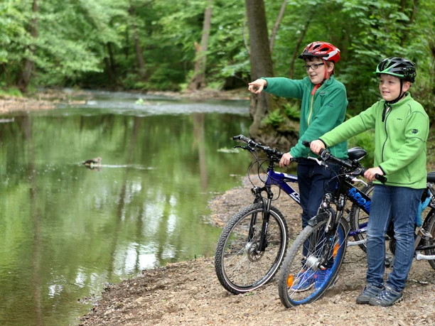 an der Dhünn Zwei Kinder mit Helmen und Fahrrädern am Flussufer, eines zeigt auf eine Ente im Wasser.
