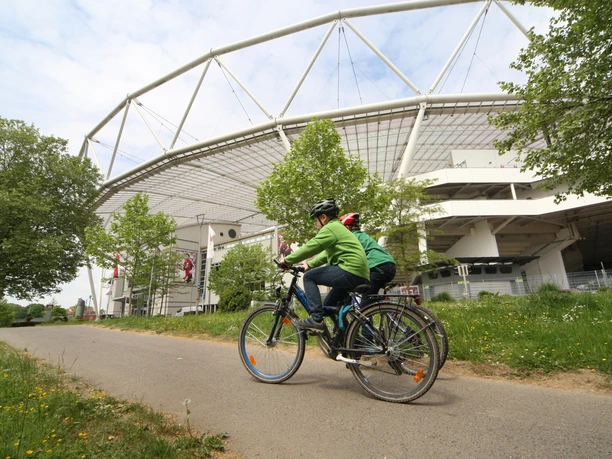 Dhünnweg an der BayArena Zwei Radfahrer fahren auf einem Weg vor einer modernen Stadionarchitektur unter blauem Himmel entlang.