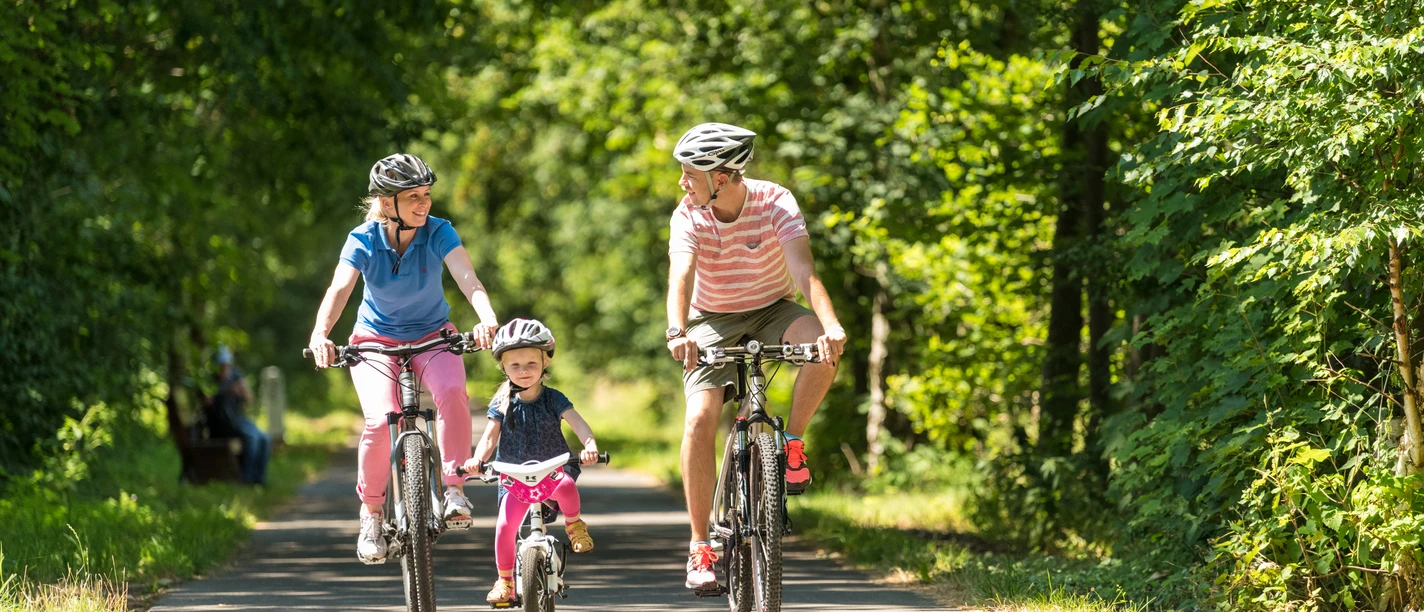 Panoramaradweg Balkantrasse Eine Familie mit einem Kind, alle mit Helmen, fährt auf einem sonnigen Waldweg Rad.