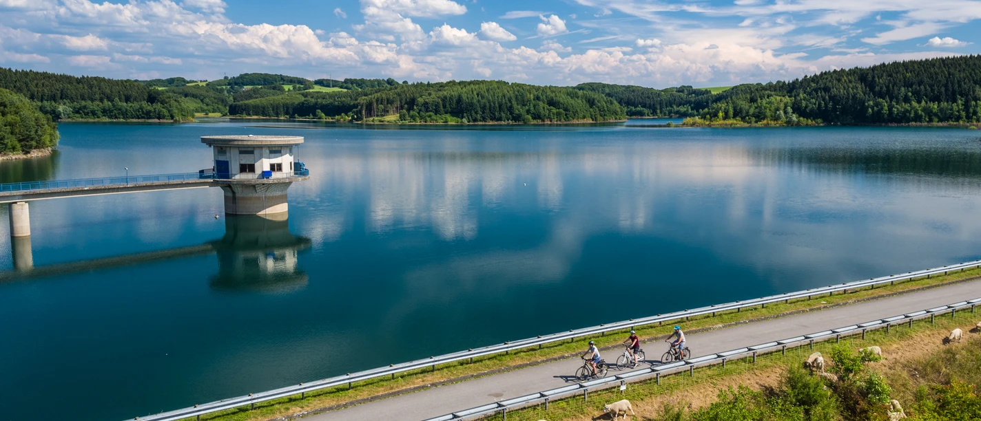 Große Dhünntalsperre Eine malerische Landschaft mit einem klaren See, Radfahrern und grünen Hügeln im Hintergrund unter blauem Himmel.
