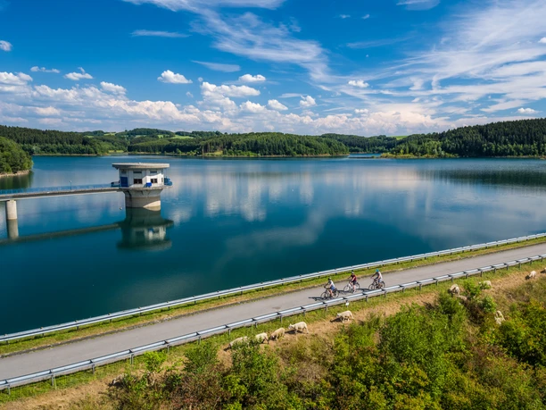 Große Dhünntalsperre Eine malerische Landschaft mit einem klaren See, Radfahrern und grünen Hügeln im Hintergrund unter blauem Himmel.