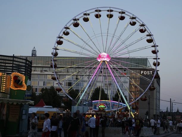 0R6A7091.JPG Riesenrad beleuchtet in Dämmerung, Menschen genießen ein Stadtfest vor modernem Gebäude.