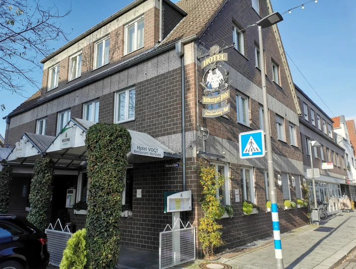 Historisches Hotel mit Ziegelsteinfassade, weißem Vordach und grünem Efeu an der Front, daneben Geschäfte.