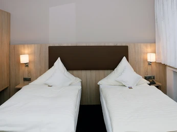 Zwei Einzelbetten mit weißen Bettdecken und Kissen in einem modern eingerichteten Hotelzimmer.