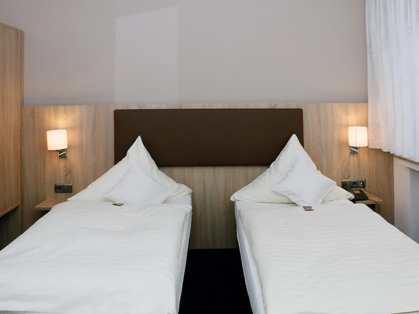 Zwei Einzelbetten mit weißen Bettdecken und Kissen in einem modern eingerichteten Hotelzimmer.