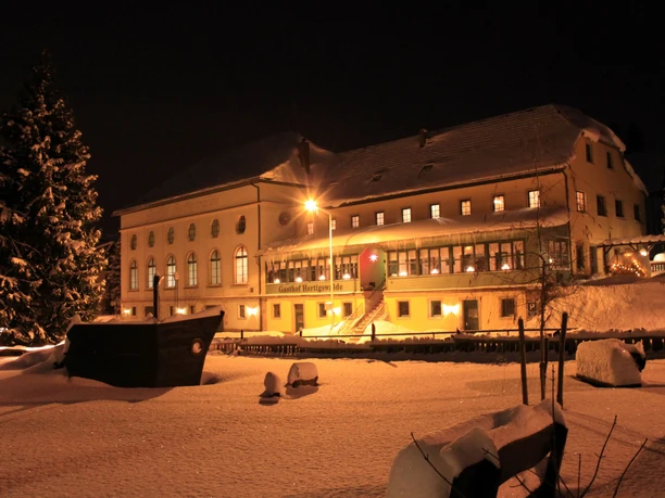 A snow-covered inn at night, illuminated by warm light, with a large, snow-covered Christmas tree in the foreground.